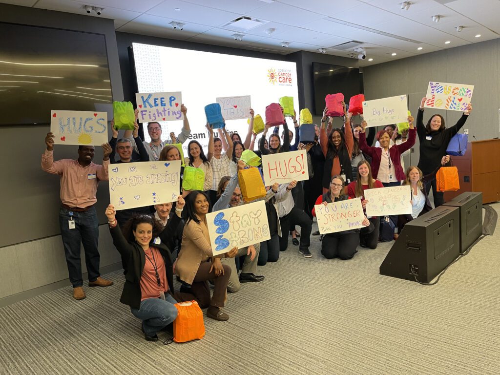 A group of people standing and kneeling in a conference room, smiling and holding up colorful posters and bags. The posters feature positive and supportive messages like 'Stay Strong,' 'HUGS,' 'You are Stronger than you Think,' and 'Keep Fighting.' Behind them is a presentation screen with the logo for 'Operation Cancer Care,' indicating this is part of a CSR team building event.