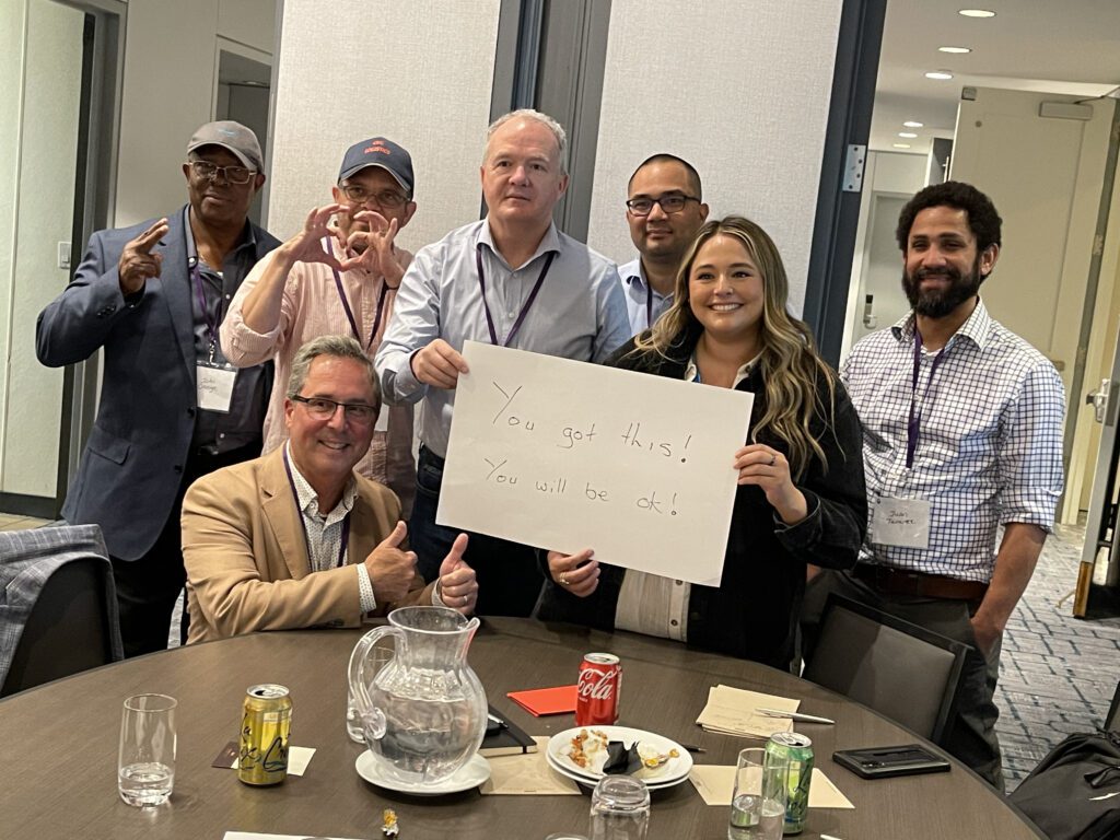 A group of seven people standing and seated around a table during a team building event. One woman holds a white sign that reads, 'You got this! You will be ok!' Others are smiling and giving thumbs up, showing supportive gestures.