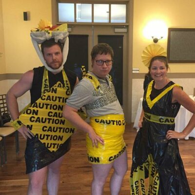 Three individuals pose confidently at a team building event, showcasing their creative outfits made from 'Caution' tape and other recyclable materials. The person on the left is wearing a black and yellow outfit, with a decorative headpiece made from caution tape. The person in the middle is also adorned in a yellow caution-tape skirt and a gray top. The individual on the right is dressed in a black and yellow dress with a matching headpiece, all made from caution tape and other materials. The mood is playful, and the group looks proud of their fashion designs.