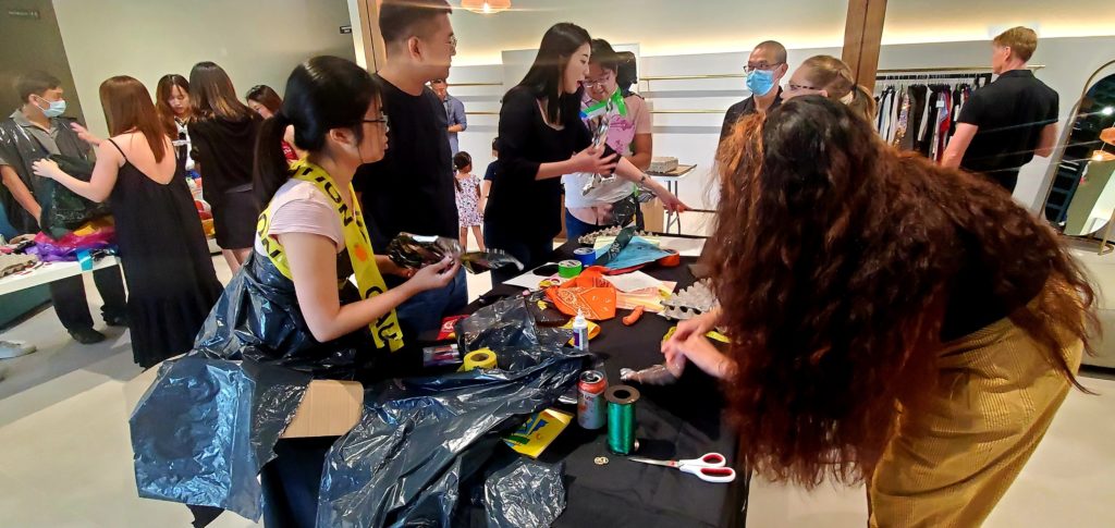 A group of participants engages in a Corporate Project Runway team building activity centered around creating fashion items. Several people are gathered around a table filled with various crafting materials such as scissors, ribbons, and recyclable items. Some participants are cutting and arranging materials while others are brainstorming or collaborating. One person wears an outfit made of trash bags and caution tape, reflecting the creative and playful nature of the event.