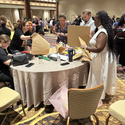 Participants at a team building event are gathered around a round table, actively engaging in a design project. They are cutting and folding pieces of cardboard and other materials as part of a creative challenge. A woman in a white dress stands next to the table, holding a piece of cardboard, while others around her are focused on their tasks. The room is filled with other groups working on similar projects.