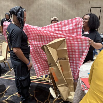 Two participants at a team building event stand, with one holding a large red and white checkered fabric, while the other appears to be modeling it or assisting in its setup. Cardboard and other recyclable materials are visible, being used to create an outfit.