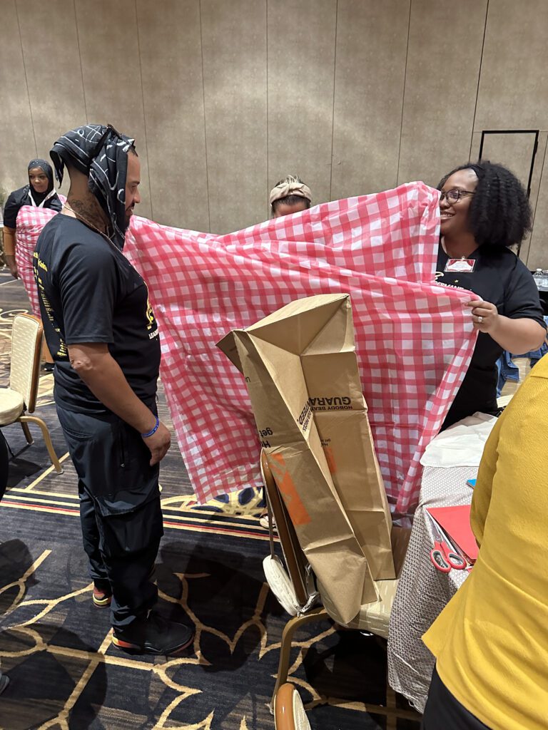 Two participants at a team building event stand, with one holding a large red and white checkered fabric, while the other appears to be modeling it or assisting in its setup. Cardboard and other recyclable materials are visible, being used to create an outfit.