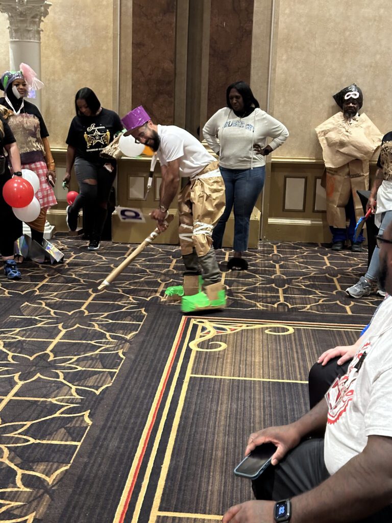 A group of people participate in a fun team building event inspired by a fashion runway show. The focus is on one individual wearing a creative outfit made from paper and other recyclable materials, including a purple hat, brown pants, and neon green shoes. The individual is in an action pose, holding a stick as if preparing for a playful activity. Several other participants, also dressed in quirky, recyclable material outfits, stand around watching.