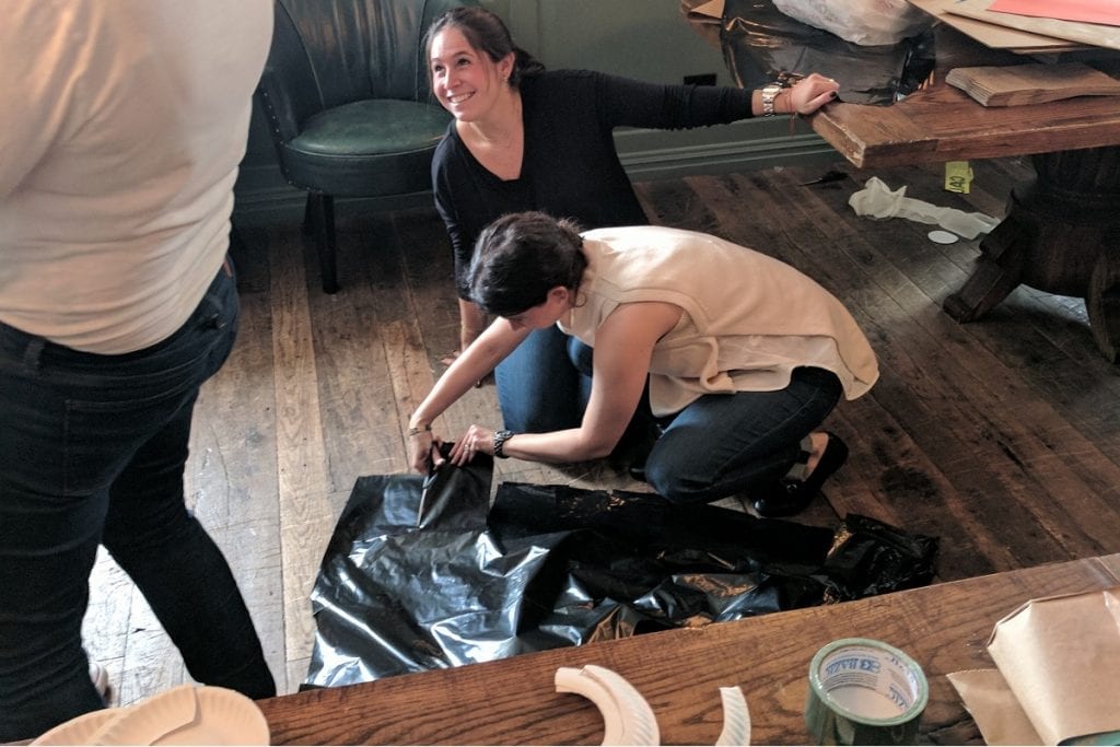 Two women engaged in a crafting activity for a team building event. One is kneeling on the wooden floor, cutting or adjusting a piece of black plastic material, while the other woman leans on a nearby table, smiling and watching.