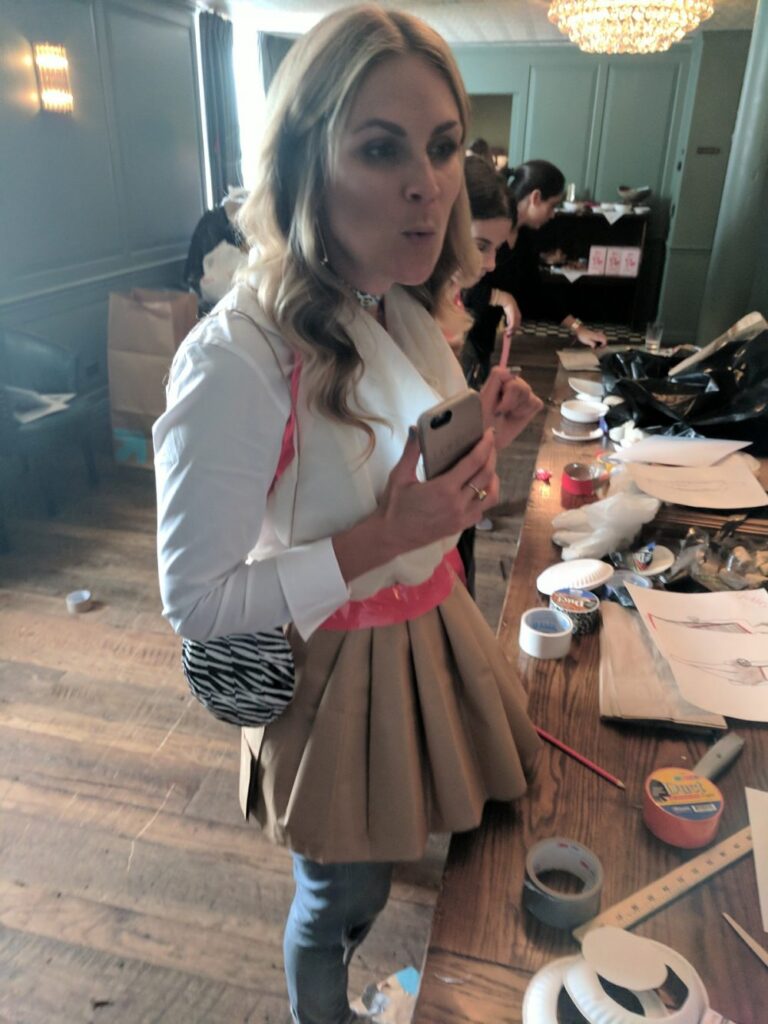 A woman standing near a table full of crafting materials, wearing a creative outfit made of paper or fabric for a team building event. Another participant is seen working in the background, focused on the materials laid out.