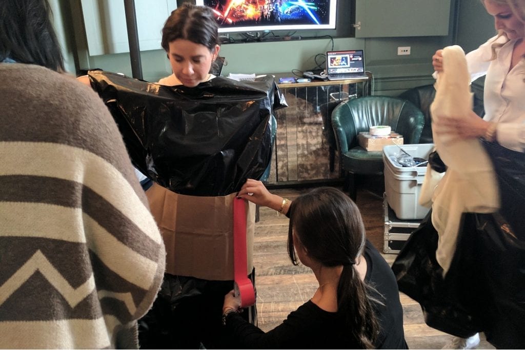 Participants collaborate during a Corporate Project Runway team building fashion activity. A woman stands in the center while others assist in constructing a garment out of black plastic material and cardboard. One person is adjusting the fit by attaching materials around her waist.