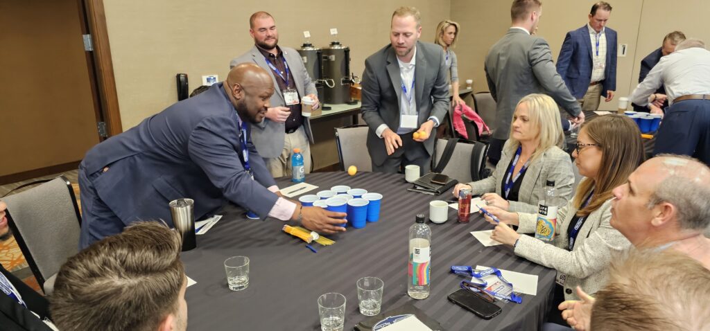 Group of professionals participating in a corporate team building event. One man arranges blue cups on the table while others watch and engage in discussion.