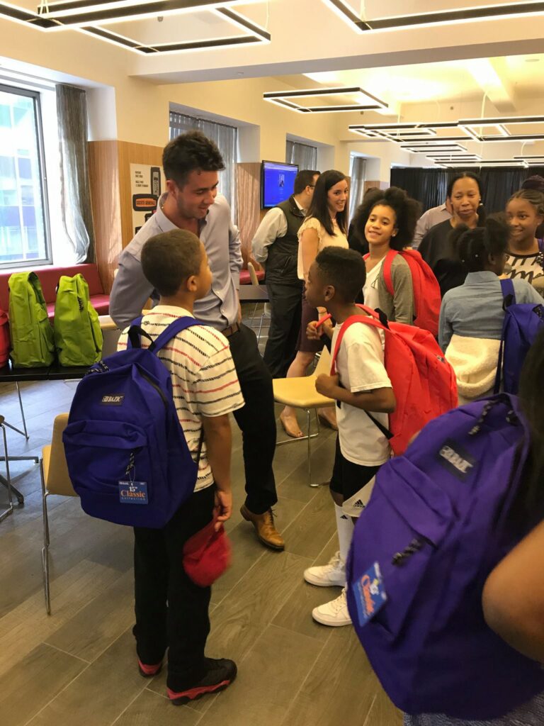 A team building participant interacts with children during a Tools for Schools CSR event, as they receive brightly colored backpacks filled with school supplies.