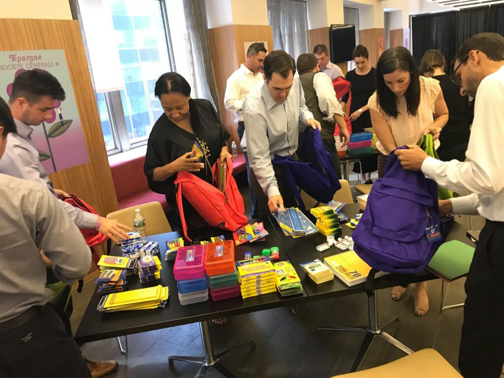 Corporate team members packing backpacks with school supplies during a csr team building event. The table is filled with notebooks, pencils, and other educational materials as participants work together to prepare donations, emphasizing teamwork and giving back to the community.