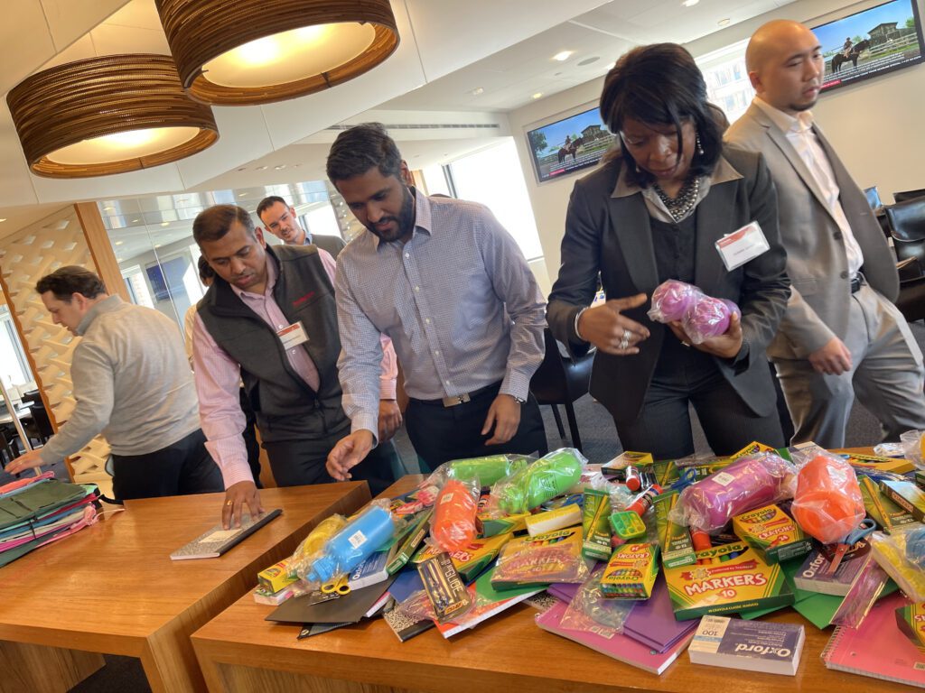 Group of professionals sorting through school supplies like markers, crayons, and notebooks on a table, preparing backpacks for donation at a Tools for Schools CSR team building event.