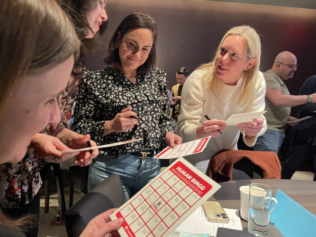 Group of colleagues engaging in a fun 'Human Bingo' team building activity, marking their bingo cards and interacting with one another.