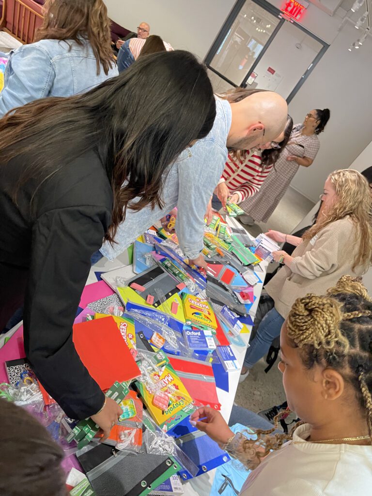 Participants at a Tools for Schools CSR event gather around a table filled with school supplies as they pack colorful folders, notebooks, and other materials into backpacks to give to students.