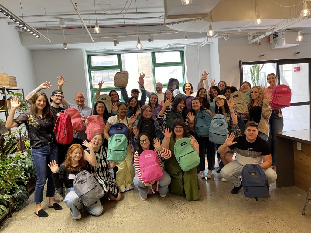 Group of enthusiastic participants holding colorful backpacks filled with school supplies, celebrating the successful completion of the Tools for Schools charity event in a bright and modern space.
