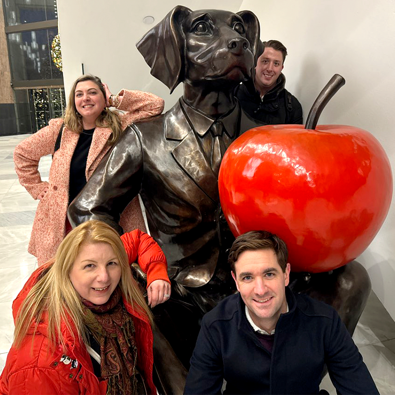 Team posing with a playful dog statue and bright red apple during the Pub Crawl Scavenger Hunt, blending exploration and friendly competition for a memorable team building experience.