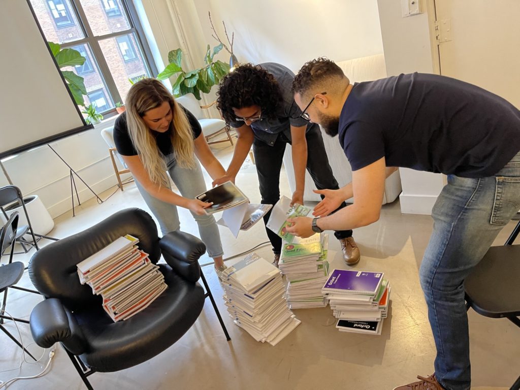 Three participants working together to organize school supplies during the 'Tools for Schools' team building csr event. They are engaged in stacking and preparing notebooks and other school materials, reflecting the event's focus on assembling backpacks for students.