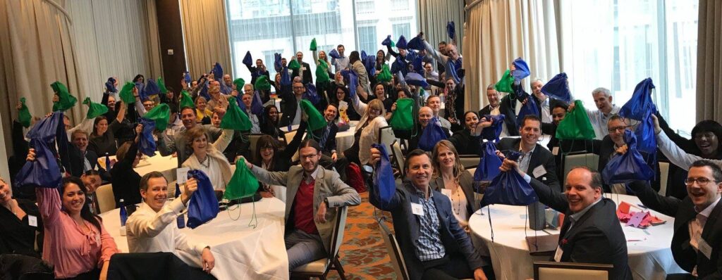 A large group of participants sits at round tables, proudly holding up green and blue drawstring bags filled with care kits during the Helping Hands team building event. The room is filled with energy as the group celebrates their collective effort to help the community.