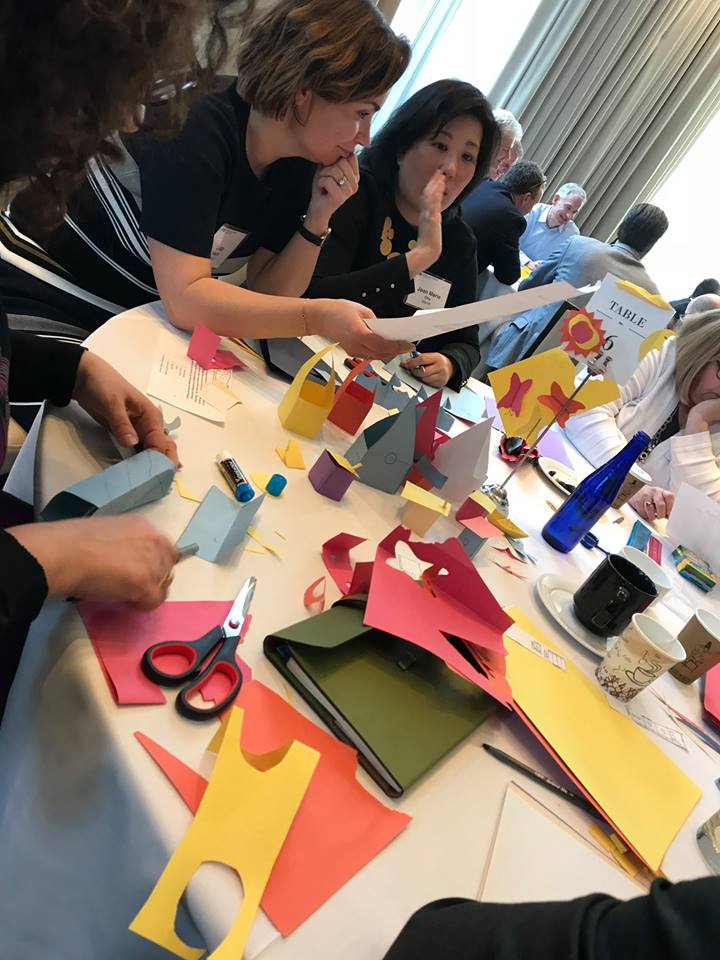 Participants work together on a creative task involving colorful paper cutouts during the Helping Hands team building event. They collaborate and discuss their progress while assembling materials for care kits.