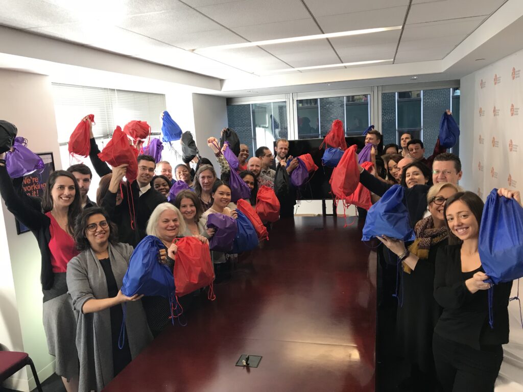 A group of participants stands around a long table, proudly holding up colorful drawstring bags filled with care kits during the Helping Hands team building event. They are smiling and celebrating their effort to support the community.
