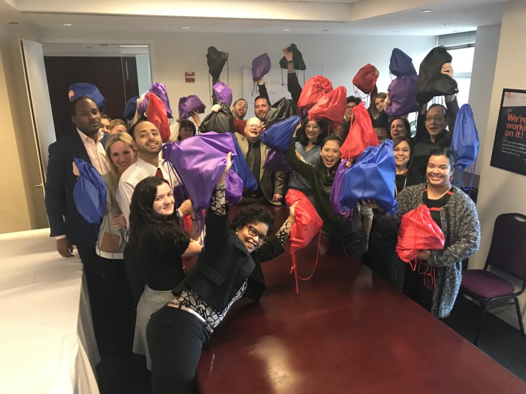 A large group of participants proudly holds up colorful drawstring bags filled with care kits they assembled during the Helping Hands team building event. The group is smiling and celebrating their contributions to the community.