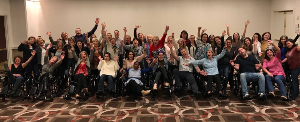 A large group of participants enthusiastically raises their hands while sitting in wheelchairs, celebrating the completion of their Wheelchairs In Motion team building event. The group is smiling and excited after successfully assembling the wheelchairs for charity.