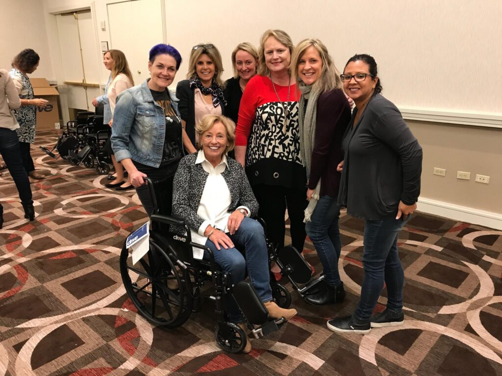 A group of participants smiles and gathers around a decorated wheelchair during the Wheelchairs In Motion team building event. One person sits in the wheelchair as the others pose together, celebrating their successful charitable effort.