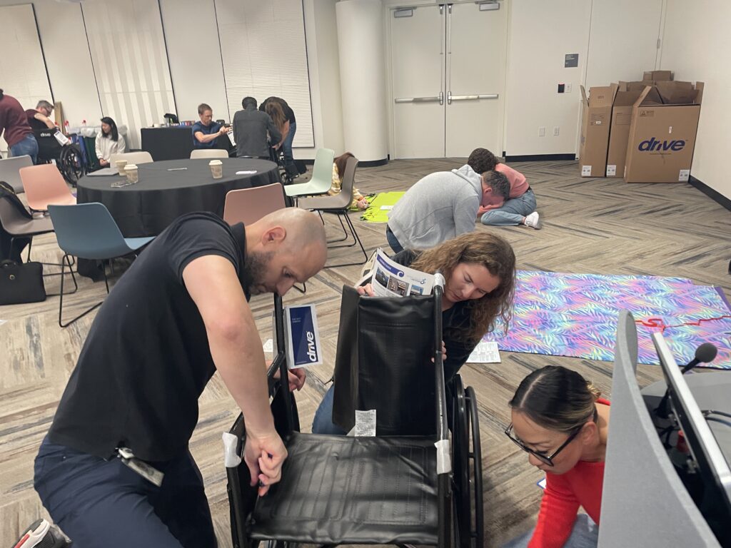 A group of participants collaborates to assemble a wheelchair during the Wheelchairs In Motion team building event. The activity is part of a charitable effort to donate wheelchairs to veterans and those in need.