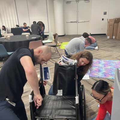 A group of participants collaborates to assemble a wheelchair during the Wheelchairs In Motion team building event. The activity is part of a charitable effort to donate wheelchairs to veterans and those in need.