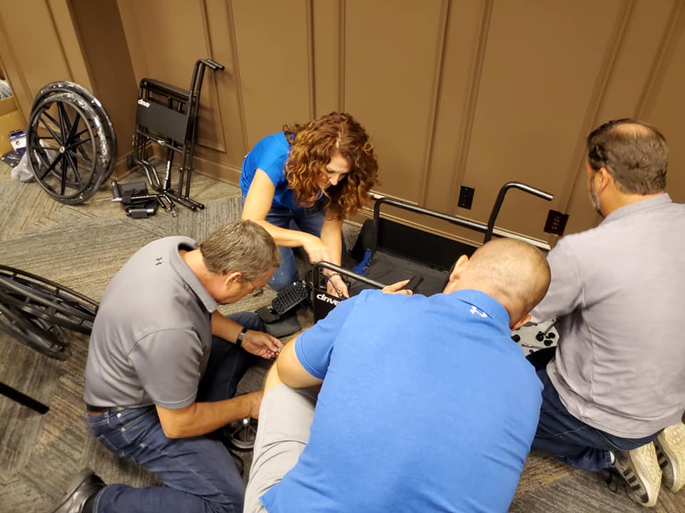 A group of participants collaborates on assembling a wheelchair during the Wheelchairs In Motion team building event. They work closely together, focusing on attaching parts to the wheelchair frame.