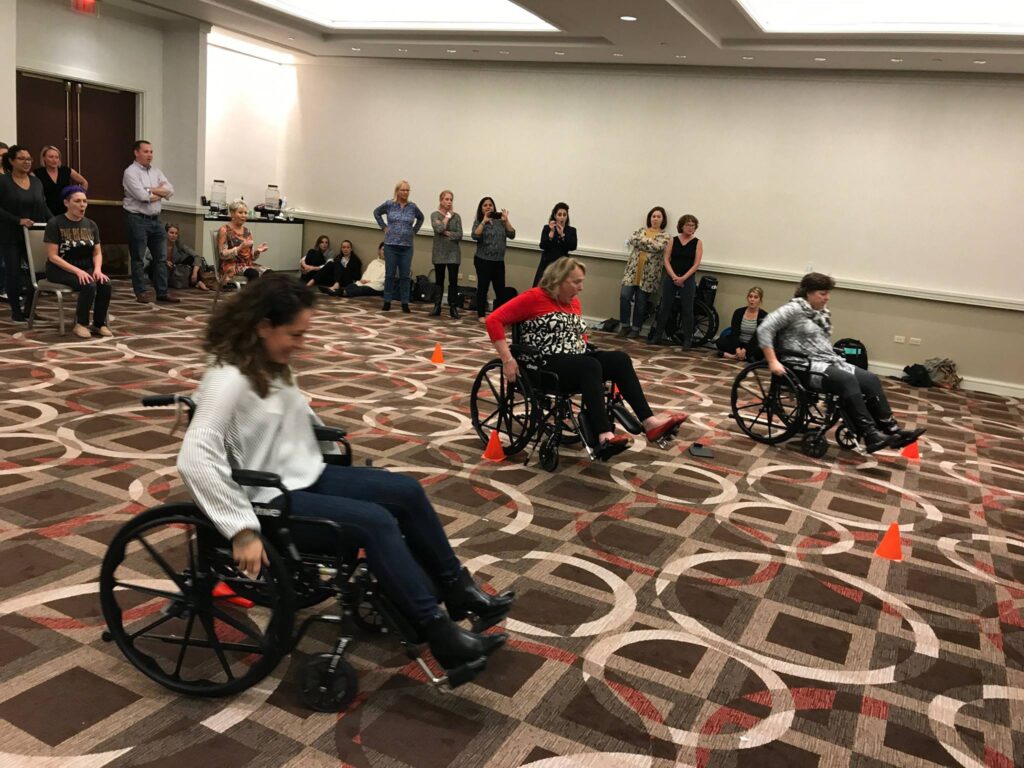 Participants race in wheelchairs through an obstacle course inside a conference room during the Wheelchairs In Motion team building event. Other attendees cheer them on as they navigate through the course.
