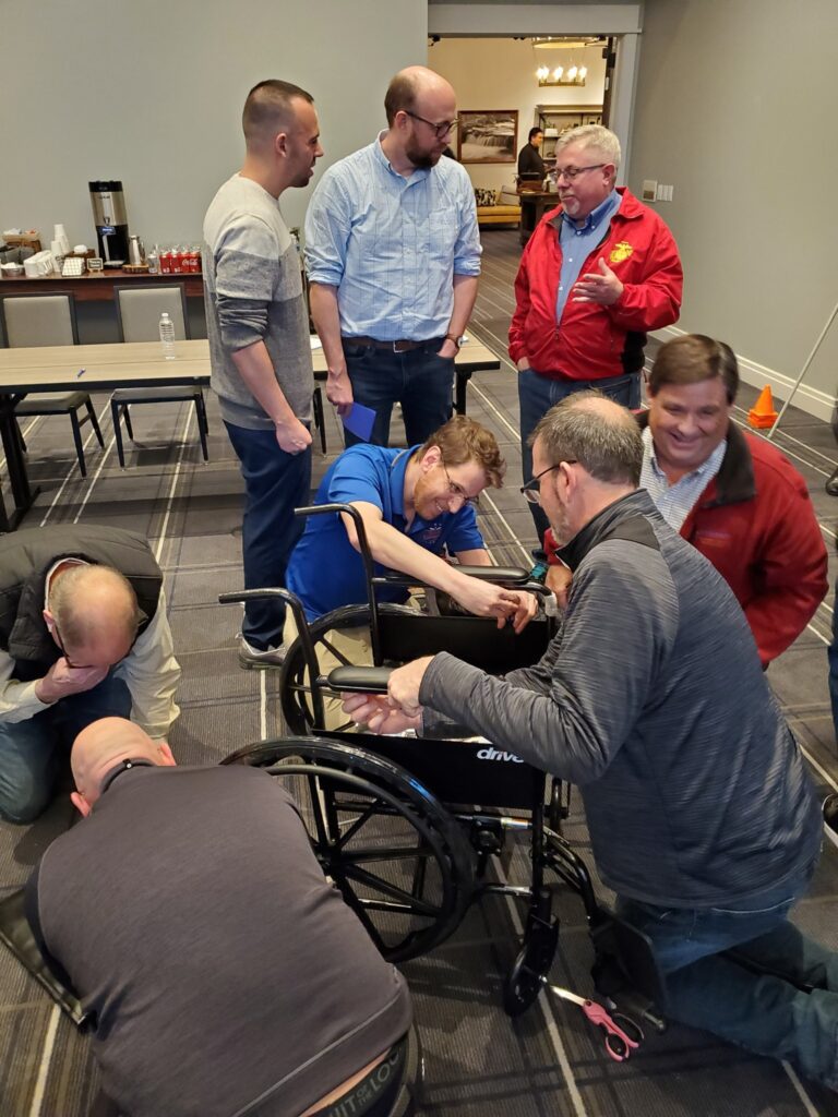 A group of participants works together to assemble a wheelchair during the Wheelchairs In Motion team building event. Several people collaborate, focusing on the task at hand, while others observe and converse in the background.