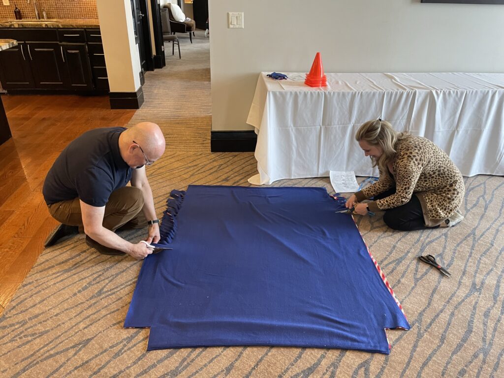 Two participants work together on the floor, cutting and preparing a large fabric piece during the Wheelchairs In Motion team building event. The fabric is part of a blanket for the wheelchair donation.
