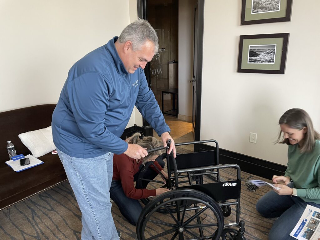 A group of participants works together to assemble a wheelchair during the Wheelchairs In Motion team building event. One participant is focused on adjusting the wheelchair, while others assist and read instructions nearby.