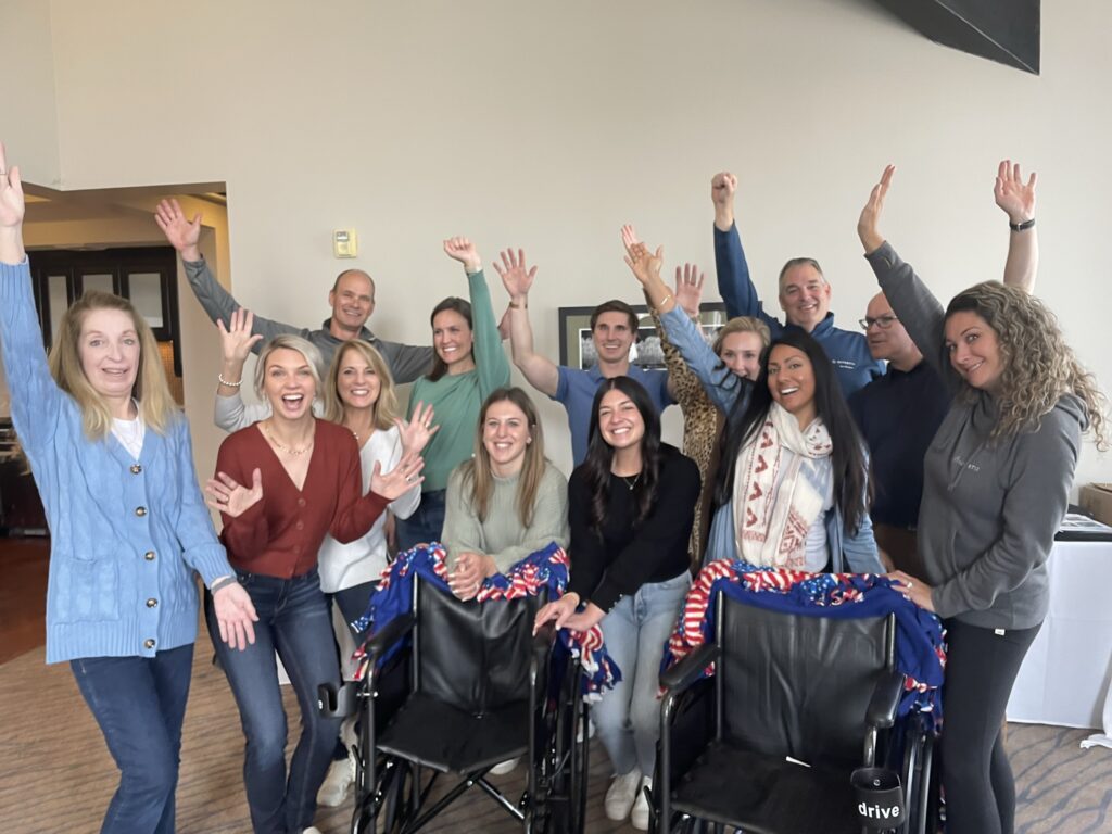 A group of participants enthusiastically raises their hands, celebrating their successful efforts during the Wheelchairs In Motion team building event. Two decorated wheelchairs are in front of the group, ready for donation.