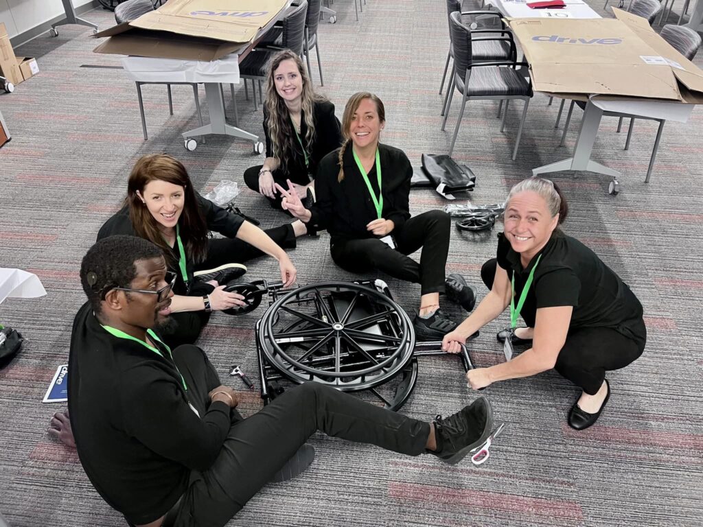 A group of smiling participants sits on the floor working together to assemble a wheelchair during the Wheelchairs In Motion team building event. The activity is part of a charitable initiative to donate wheelchairs to those in need.