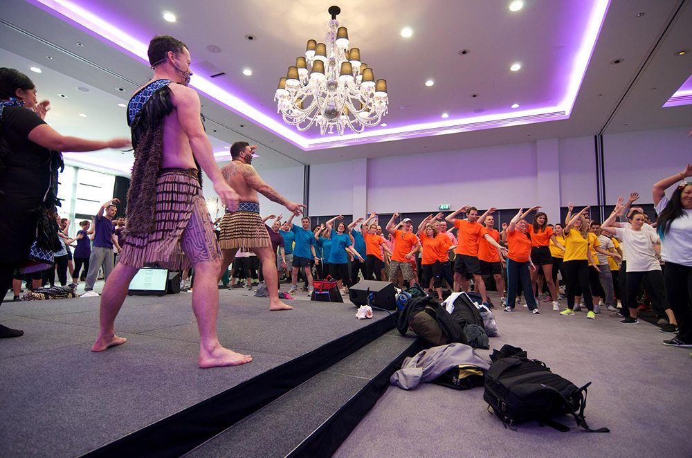 Maori performers in traditional attire leading a Haka team building session for a large group of participants.