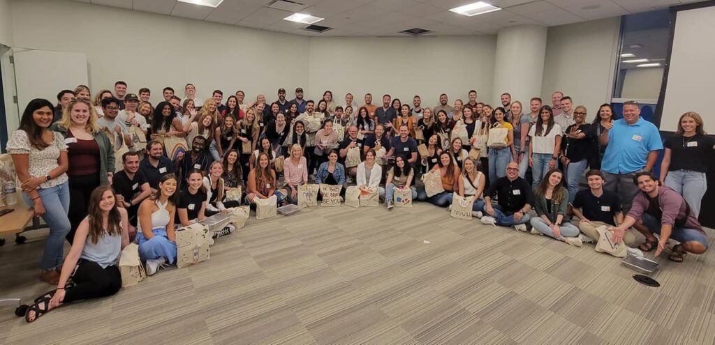 A large group of participants pose for a group photo at the conclusion of a team building cooking activity. The group, consisting of over 50 people, is smiling and seated on the floor of a spacious room. Many participants are holding decorated bags, filled with food donations they prepared during the event. The atmosphere appears to be joyful and celebratory, marking the success of their collaborative efforts to give back to the community.