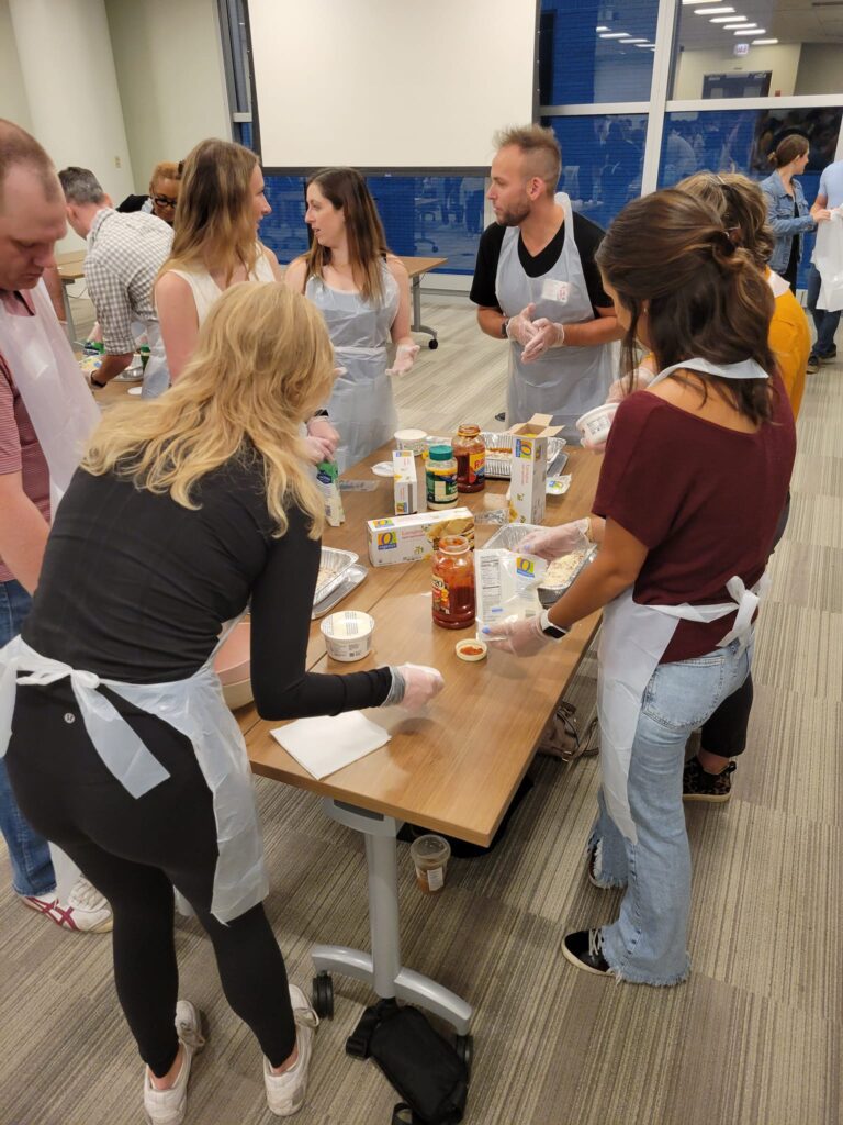 A group of participants wearing aprons and gloves are engaged in a team building cooking activity. They are gathered around a table, preparing ingredients like pasta sauce and cheese to assemble lasagna dishes. The group is conversing and collaborating, with a variety of food items and kitchen tools spread out on the table. The setting appears lively and focused as they work together to prepare meals for donation.