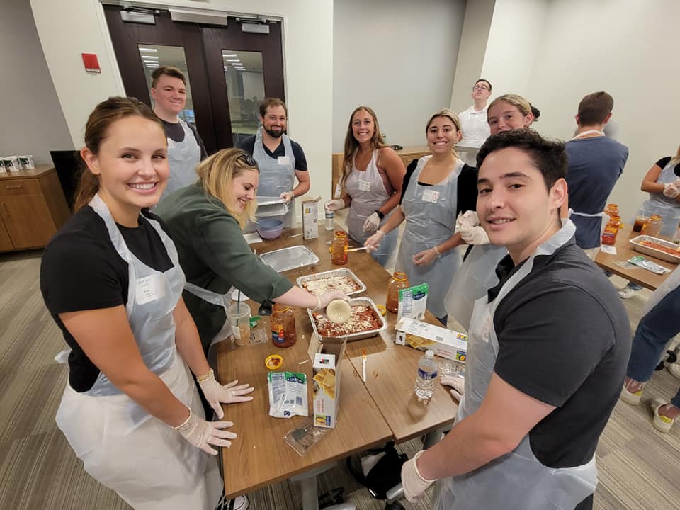 A group of nine individuals is gathered around a table, all smiling and wearing aprons and gloves as they participate in a team building cooking activity. One person is scooping cheese onto a lasagna dish, while various other ingredients like sauce, pasta, and cheese are arranged on the table. The group looks happy and engaged in their task as they prepare meals together.