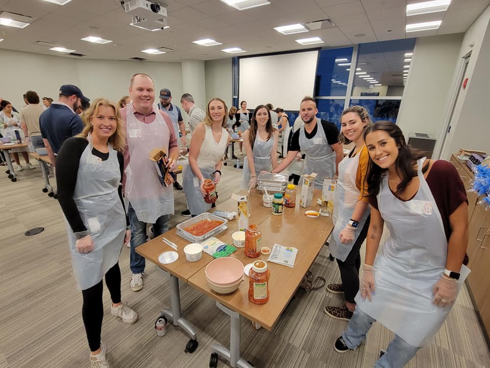 A group of eight people stands around a table, smiling for the camera while participating in a team building cooking activity. Each person is wearing an apron and gloves, and various ingredients such as pasta, sauce, and cheese are spread out on the table. Some of the participants are holding jars of sauce, ready to assemble a dish. The group is engaged in preparing meals as part of a community service project.