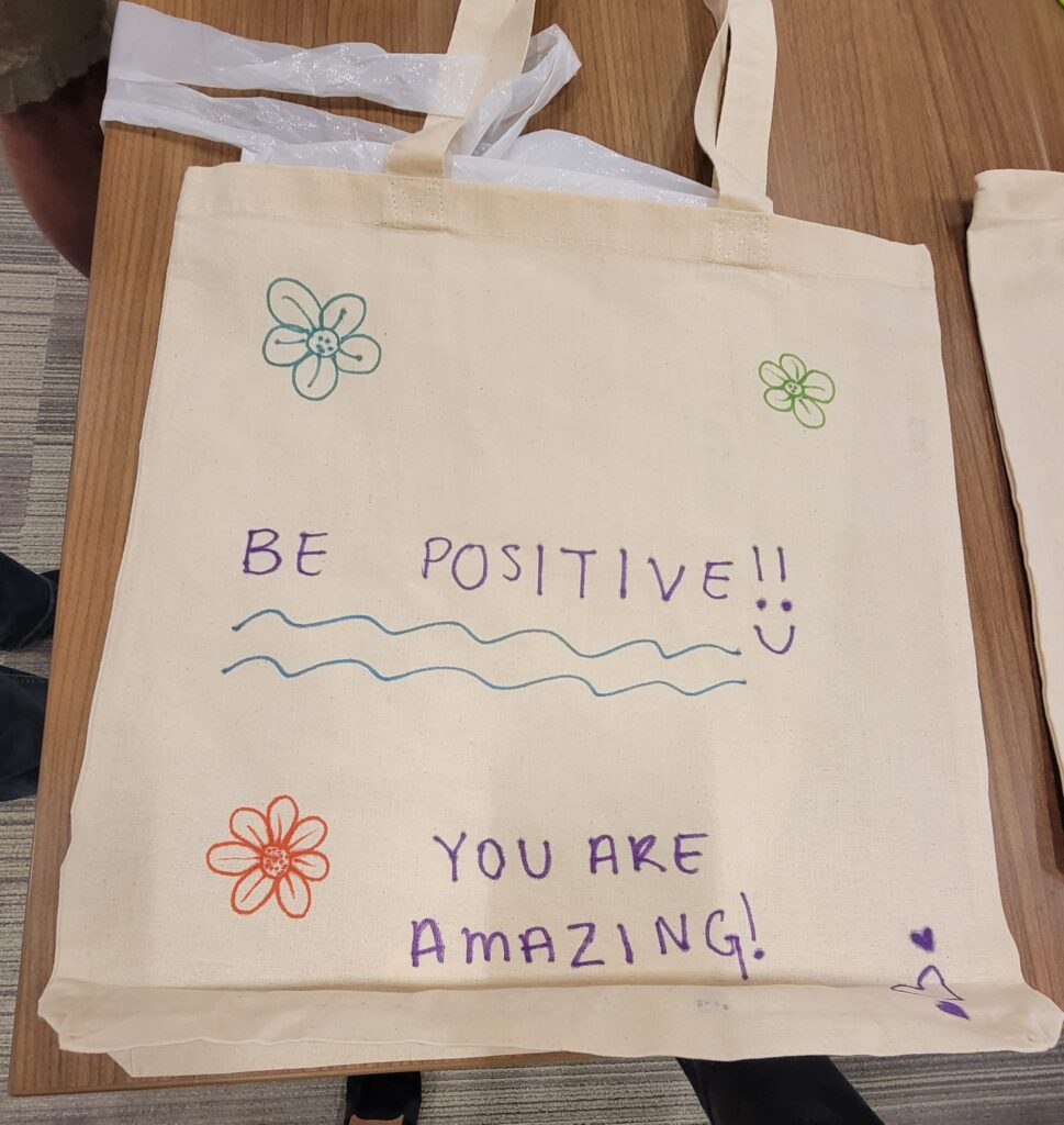 A decorated canvas tote bag lays flat on a wooden table. The bag features hand-drawn flowers in various colors and motivational phrases written in colorful markers. The text on the bag reads 'BE POSITIVE!!' with a smiley face, followed by 'YOU ARE AMAZING!' below. The design is simple and cheerful, meant to inspire and encourage.