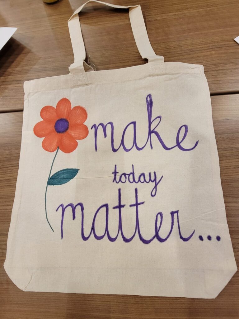 A beige fabric tote bag displayed on a wooden table, decorated with a hand-painted orange flower with a purple center and green stem. Next to the flower, the words 'make today matter...' are written in purple cursive, encouraging a positive message. The bag is part of a CSR team building event.