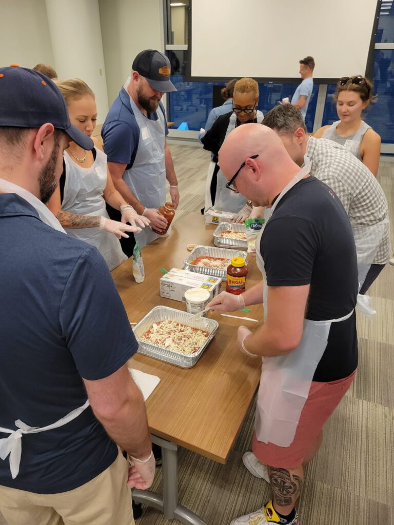 A group of participants wearing aprons and gloves are gathered around a table, preparing trays of pasta for donation. They are adding ingredients like sauce and cheese to large aluminum trays. The participants are engaged and working together in a well-lit room, with various ingredients and supplies on the table. This is part of a CSR team building event aimed at preparing meals for those in need.