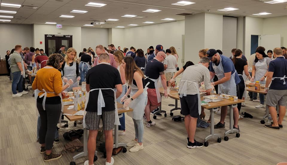 A large group of participants, all wearing aprons, are spread across multiple tables in a spacious room. They are actively preparing food together, working in small teams to assemble meals for donation. The room is well-lit, with several rows of tables and individuals focused on their tasks, contributing to a community service cooking event aimed at making food for those in need.