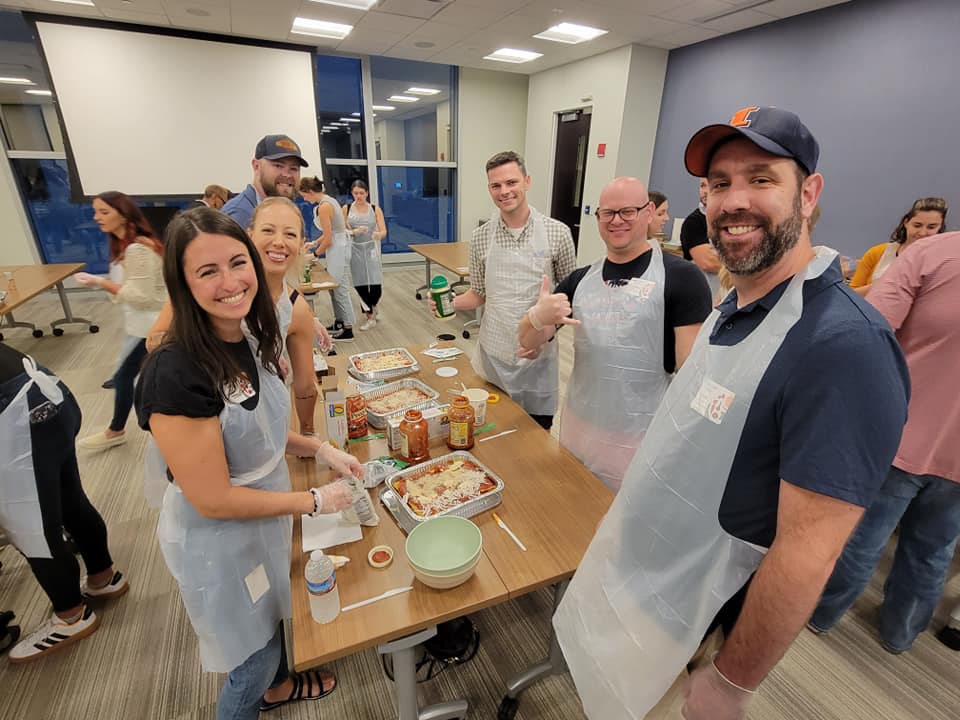 A group of six people is gathered around a table during a team building cooking activity, smiling and wearing aprons and gloves. They are preparing meals, with lasagna trays and various ingredients like pasta, sauce, and cheese laid out on the table. One member gives a thumbs-up to the camera, and the group looks cheerful and engaged in the activity.