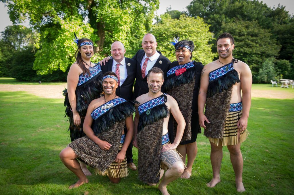 Group photo of Maori performers in traditional attire alongside two suited participants during a Haka team building event in a scenic outdoor setting.