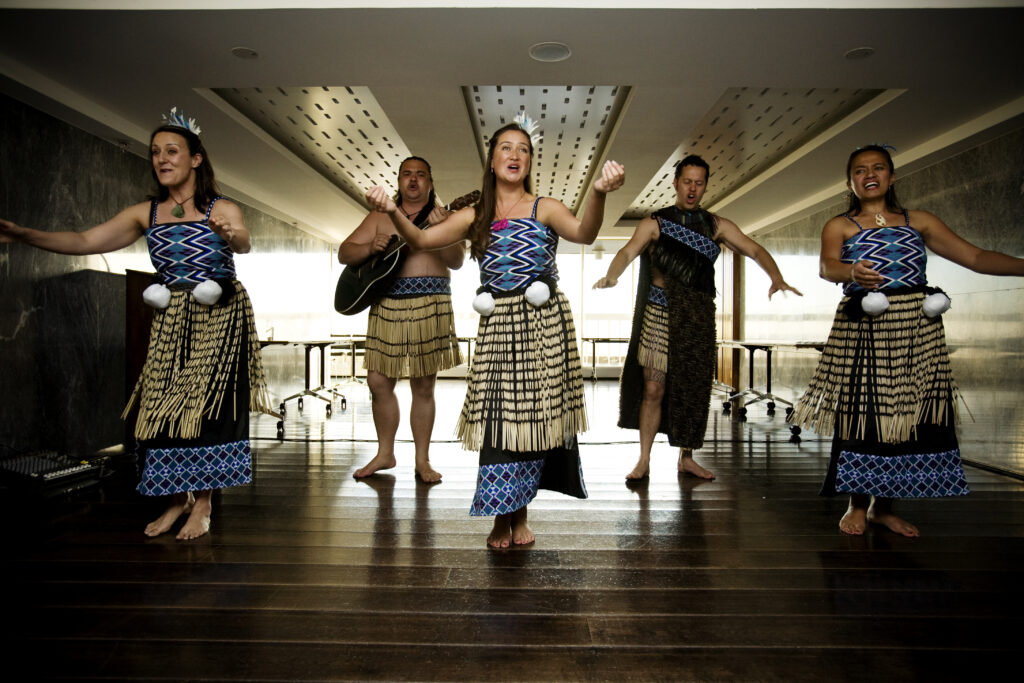 Maori performers in traditional attire deliver an energetic Haka team building experience indoors, showcasing dance, chant, and cultural instruments.