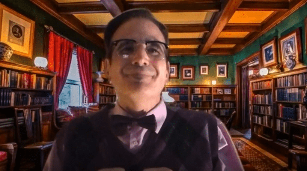 A man wearing glasses, a bow tie, and a patterned sweater vest is seated in a library setting with shelves filled with books behind him. The man is in character, part of a role-playing murder mystery team building event.