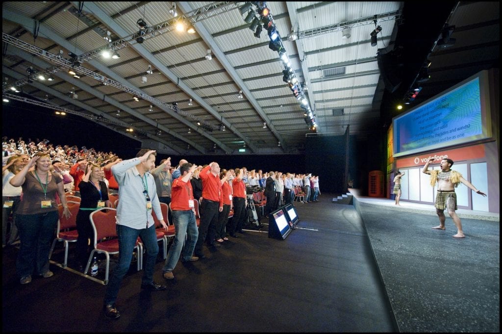 Large audience in a conference hall actively participating in a Haka team building event led by Maori performers on stage.