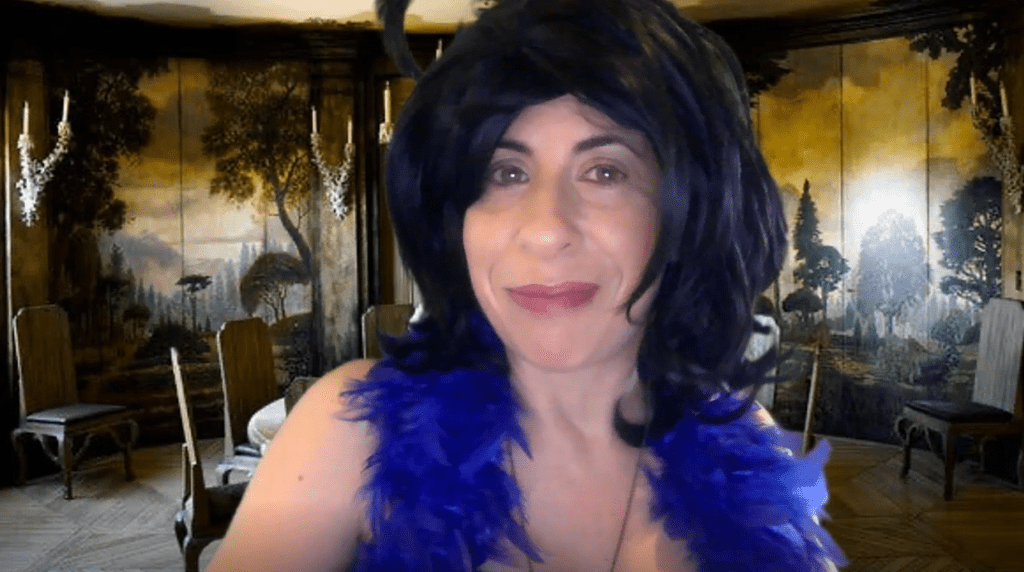 A woman with a playful expression, wearing a black wig and a bright blue feather boa, poses in front of a luxurious room decorated with an artistic mural of trees and a grand dining table. The setting evokes a sense of elegance and mystery. She is playing a role in a themed murder mystery team building event.